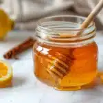 Asian honey protocol recipe in open glass jar with turmeric honey blend, honey dipper, lemon and cinnamon on white marble