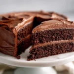 moist vegan chocolate cake slice with thick chocolate frosting on white ceramic cake stand