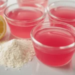 Four glass ramekins filled with firm glossy pink dr gupta jelly fit gelatin on a white countertop with halved lemon and protein powder beside them