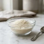 Bovine gelatin powder in a clear glass bowl with a spoon on a white surface.
